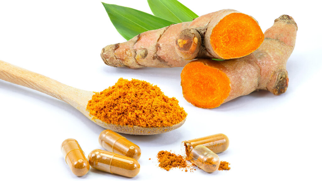 Racines de curcuma coupées avec des capsules de supplément et de la poudre de curcuma sur une cuillère en bois.