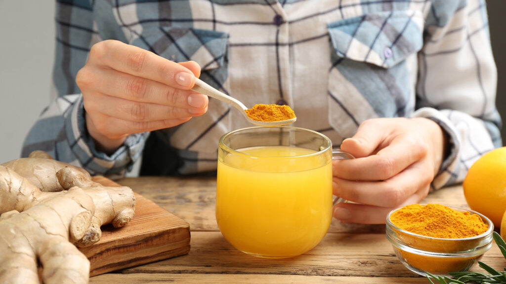 Mains préparant une boisson au curcuma avec une cuillère de poudre de curcuma ajoutée à un verre de boisson jaune.