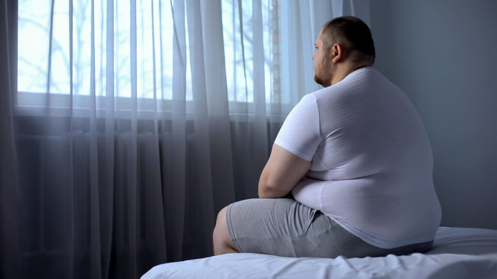 Homme assis sur un lit regardant par la fenêtre, illustrant l'isolement lié à l'obésité.