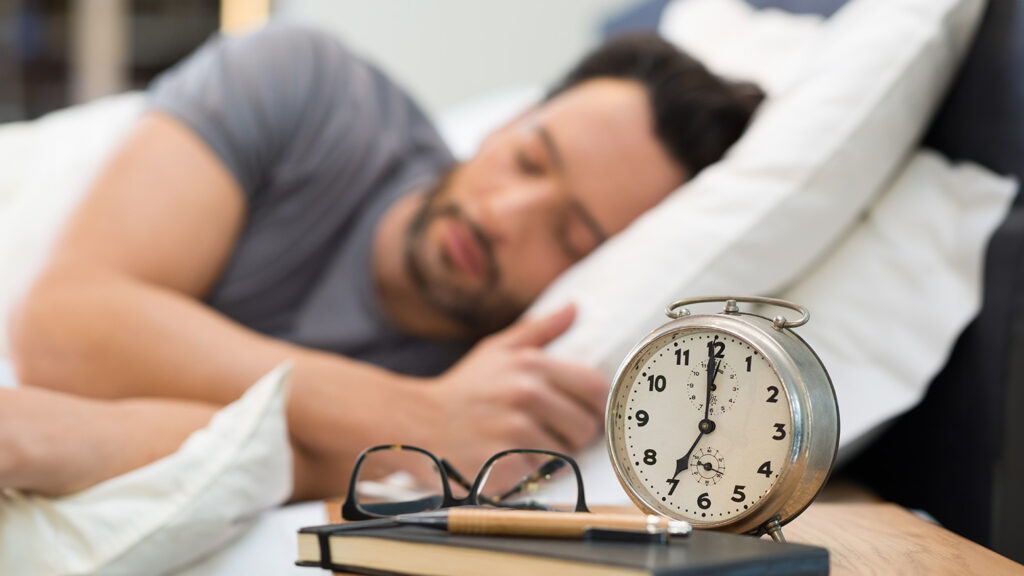 Un homme endormi dans son lit avec un réveil à l'avant-plan indiquant qu'il est l'heure de se lever.