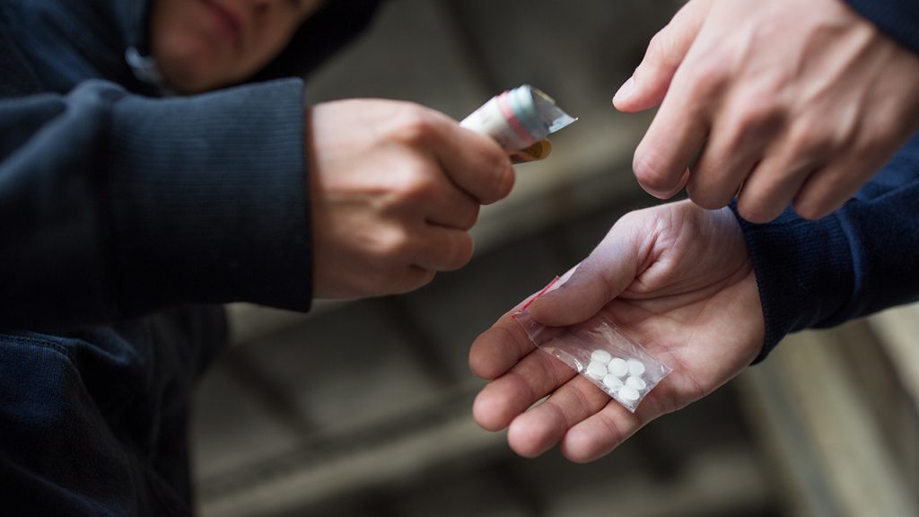 Transaction de drogue entre deux personnes, une donnant des comprimés dans un sachet plastique et l'autre tenant des billets.