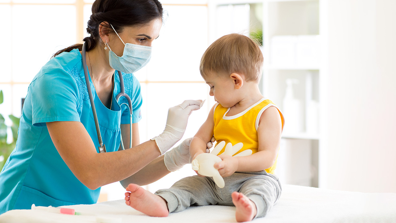 Un médecin en blouse bleue vacciné un nourrisson assis sur une table.