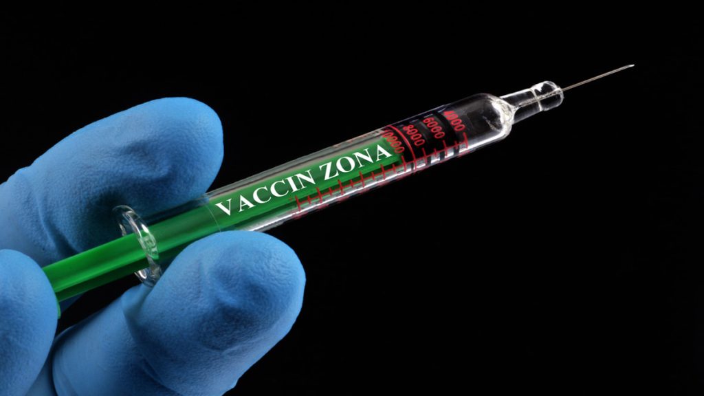 Seringue avec l'inscription 'Vaccin Zona', tenue par une main gantée.